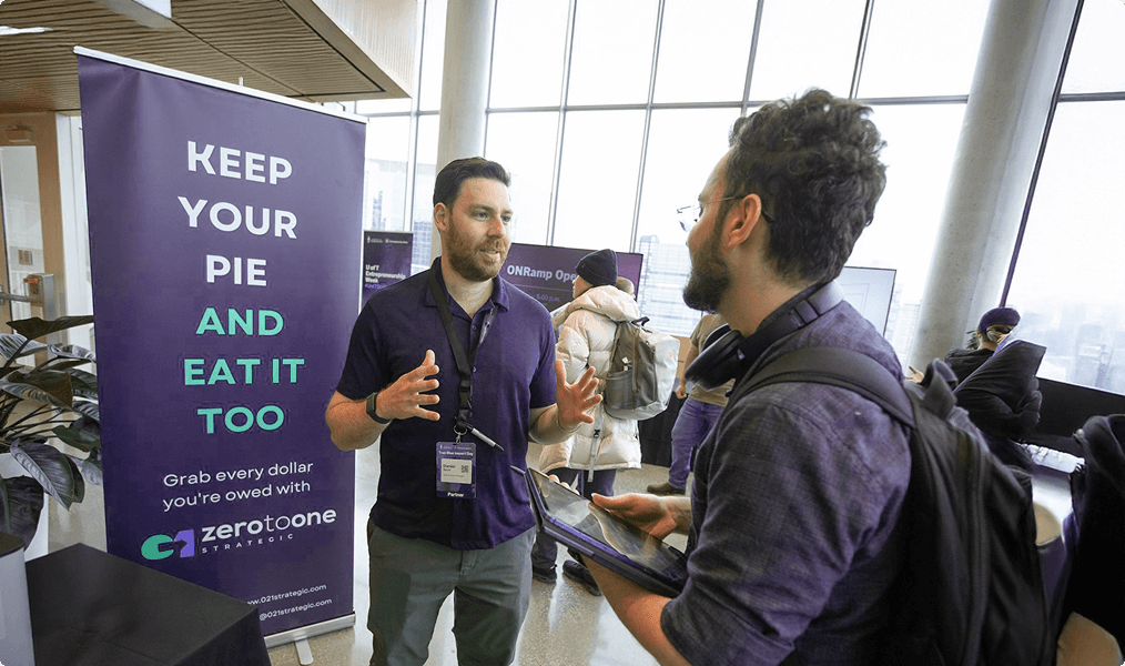 A picture of Dan talking to a potential client at an event in front of a Zero to One banner.